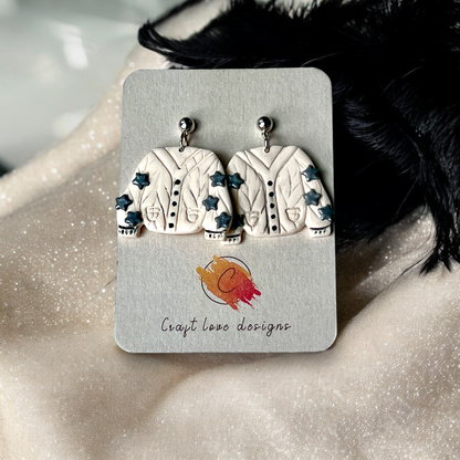 Quirky sweater-shaped drop earrings in cream with blue star accents, displayed on a card against a soft beige, sparkly fabric backdrop. 