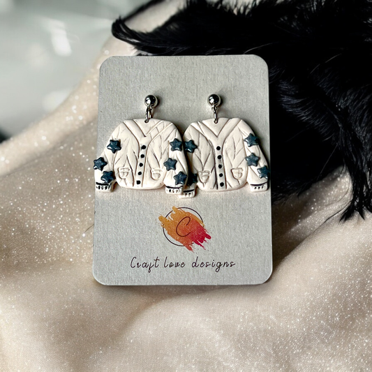 Quirky sweater-shaped drop earrings in cream with blue star accents, displayed on a card against a soft beige, sparkly fabric backdrop. 