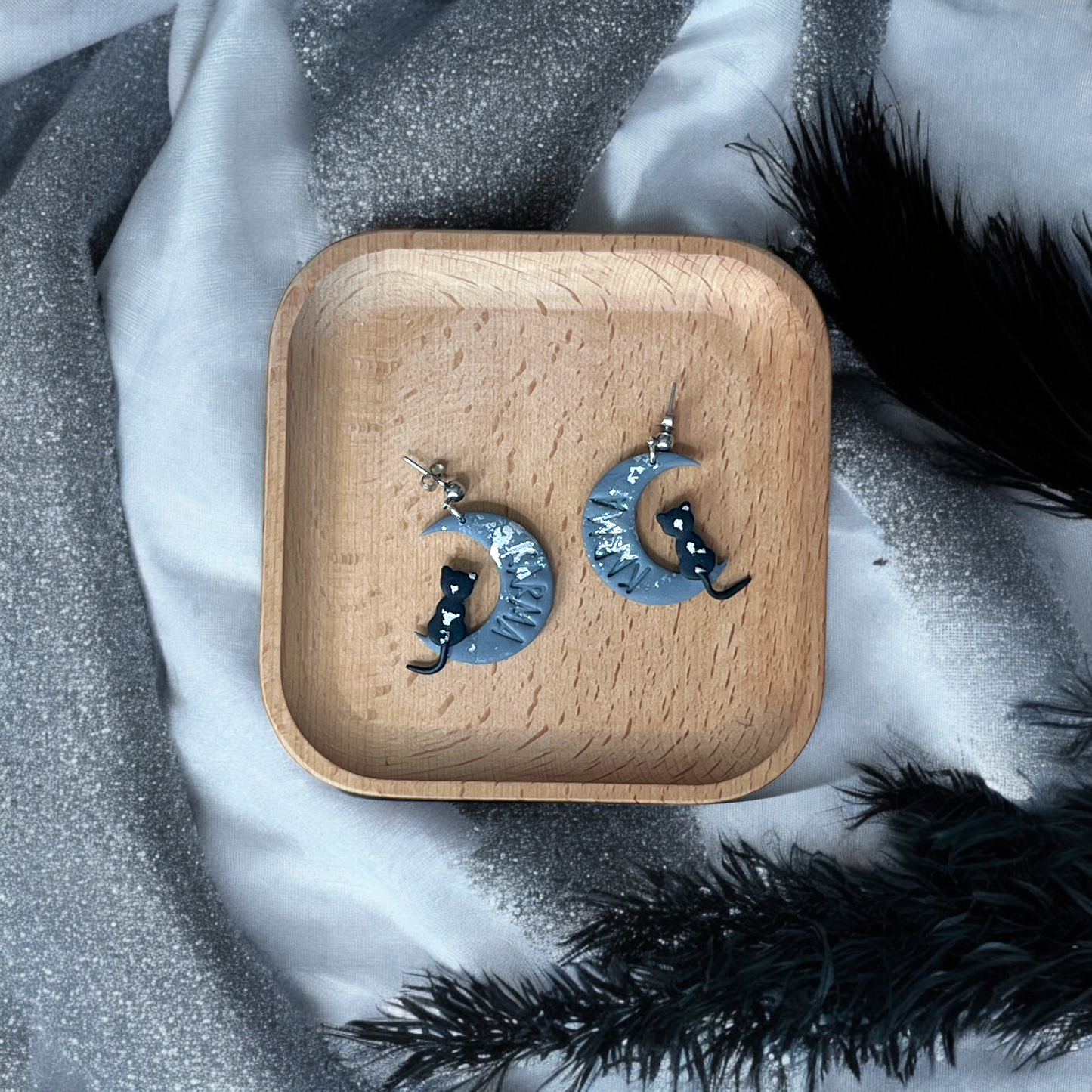 Crescent moon earrings with black cats and silver accents, placed on a wooden tray surrounded by soft, sparkly silver and black fabrics.