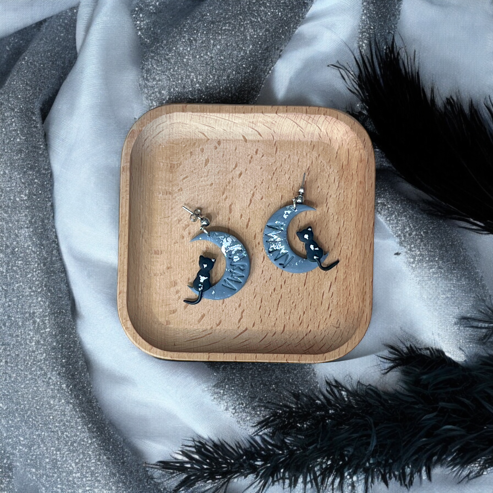 Crescent moon earrings with black cats and silver accents, placed on a wooden tray surrounded by soft, sparkly silver and black fabrics.