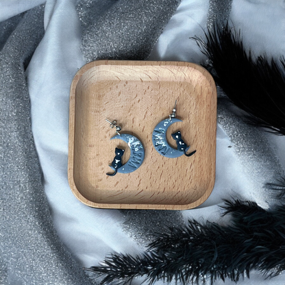 Crescent moon earrings with black cats and silver accents, placed on a wooden tray surrounded by soft, sparkly silver and black fabrics.