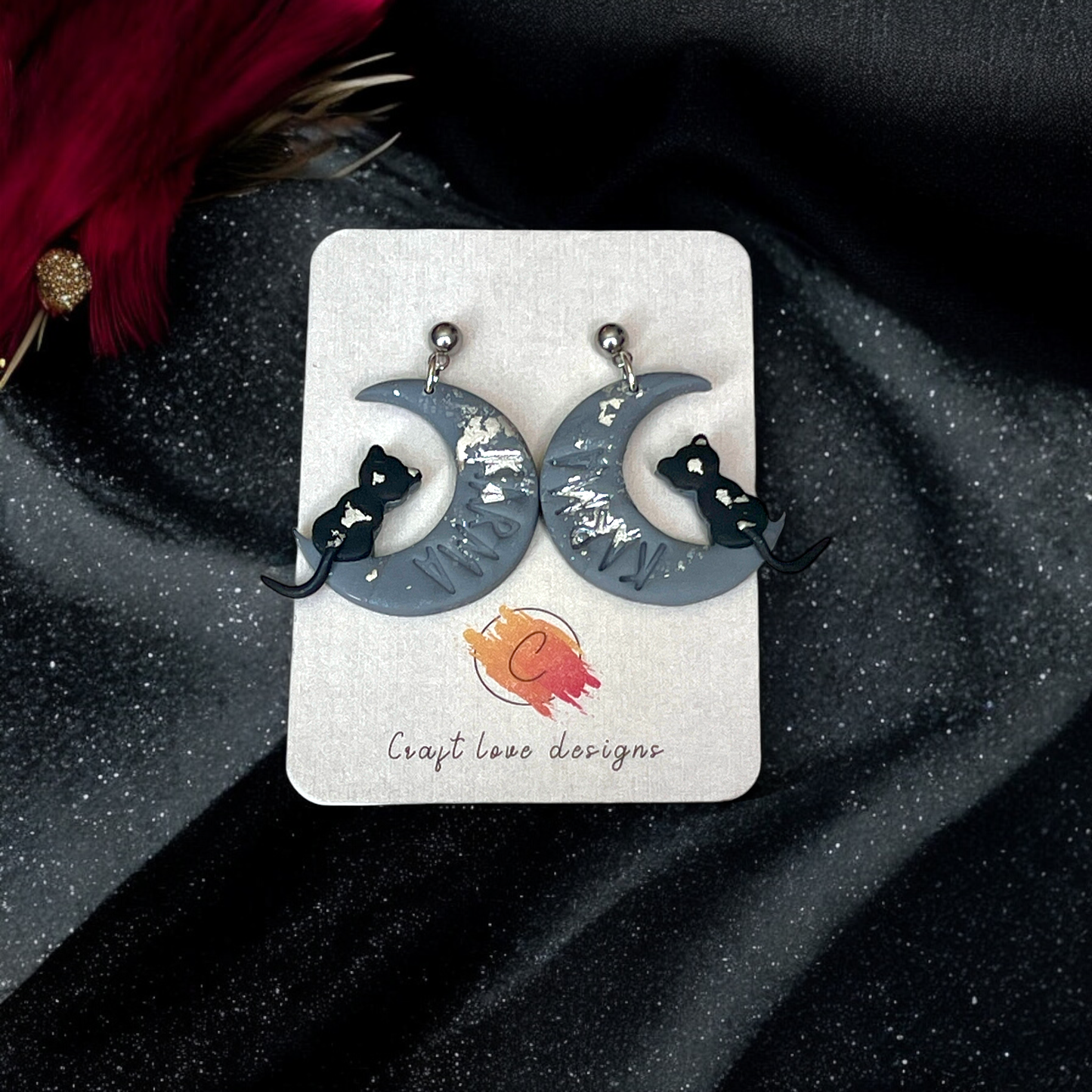 Crescent moon earrings featuring black cat silhouettes and silver accents, displayed on a Craft Love Designs card with a glittery black background.