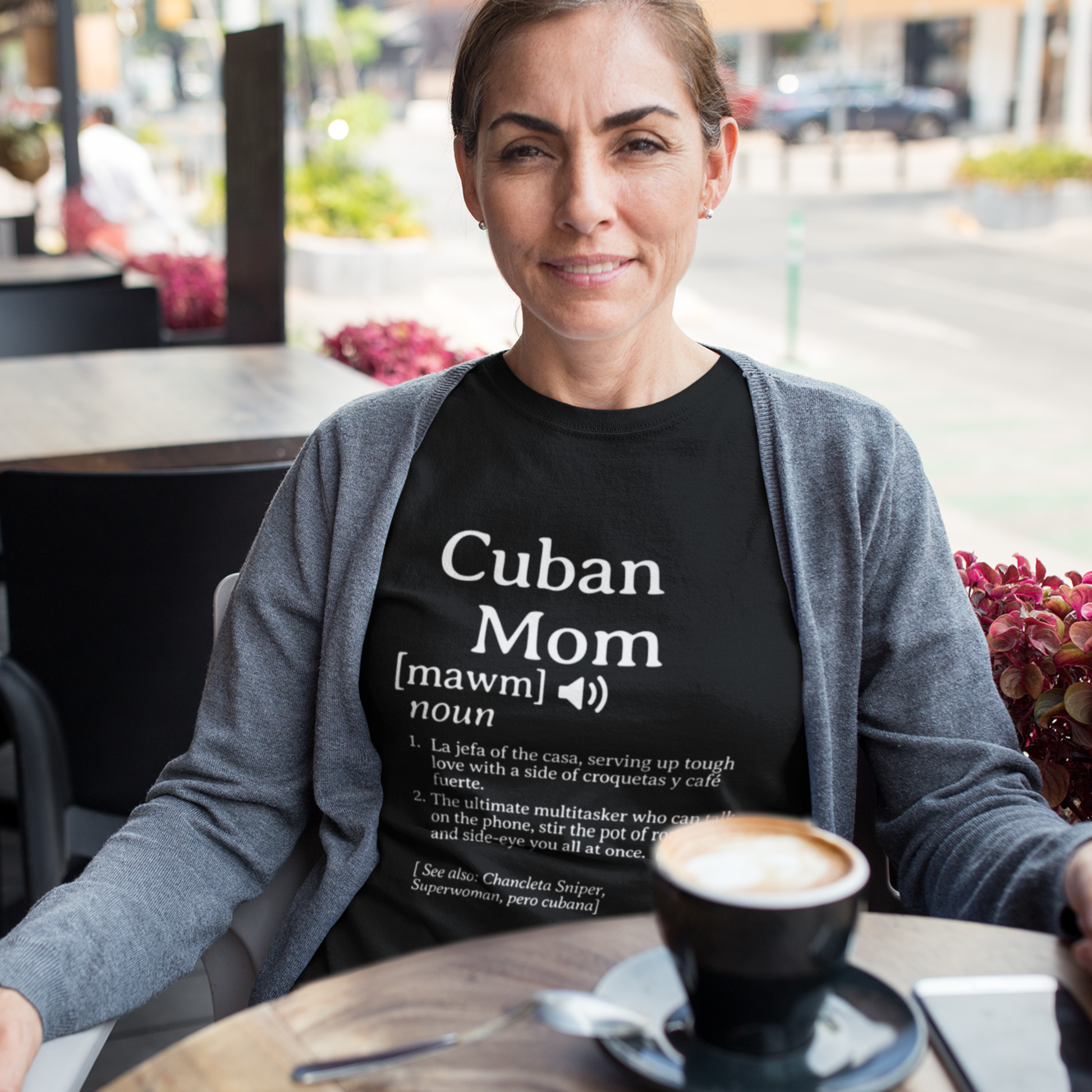Woman wearing a Cuban Mom definition shirt, sitting at a café table with a cup of coffee and a casual, relaxed look.