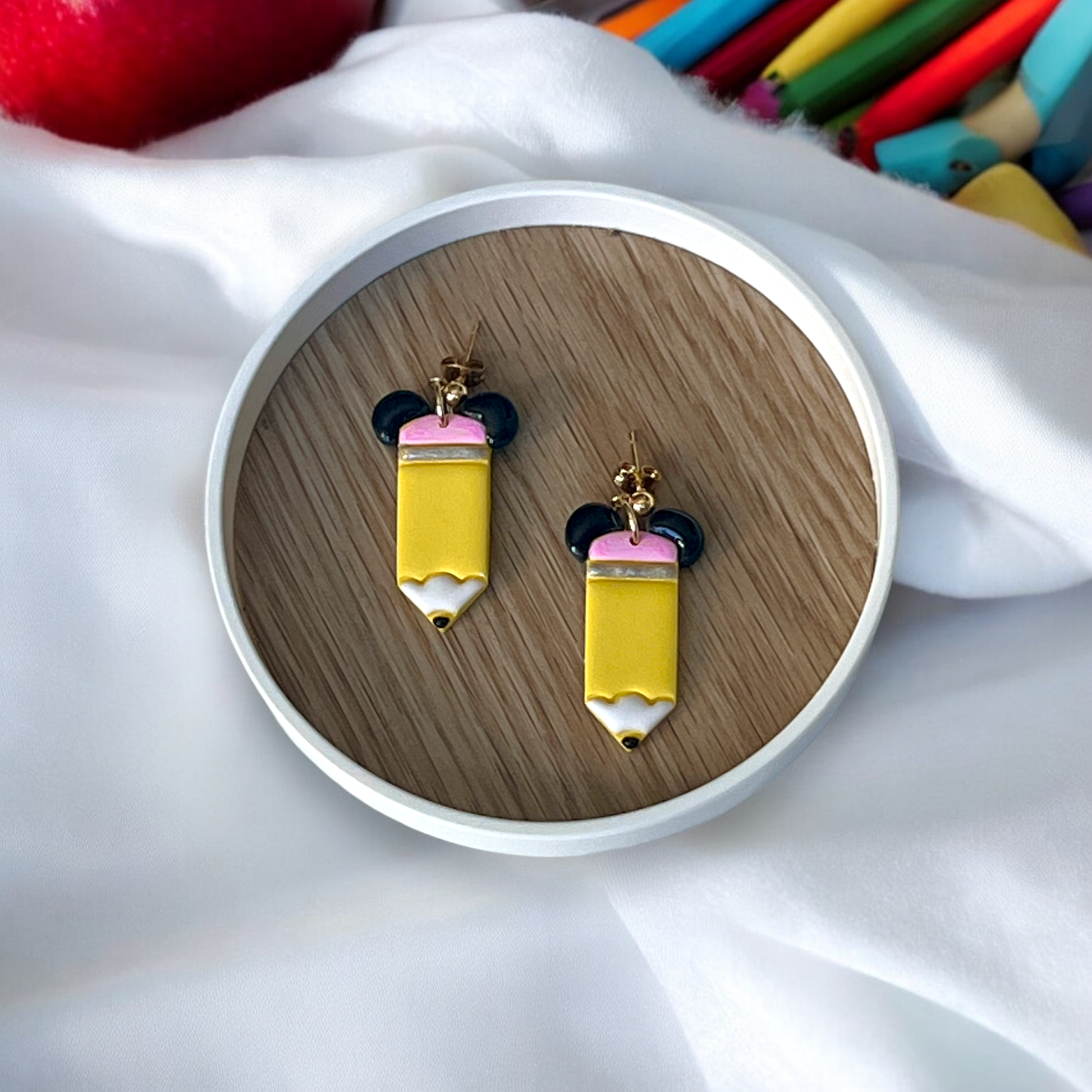 Handmade yellow pencil earrings with black ear accents, placed in a round wooden dish over white fabric and colorful scattered pencils.