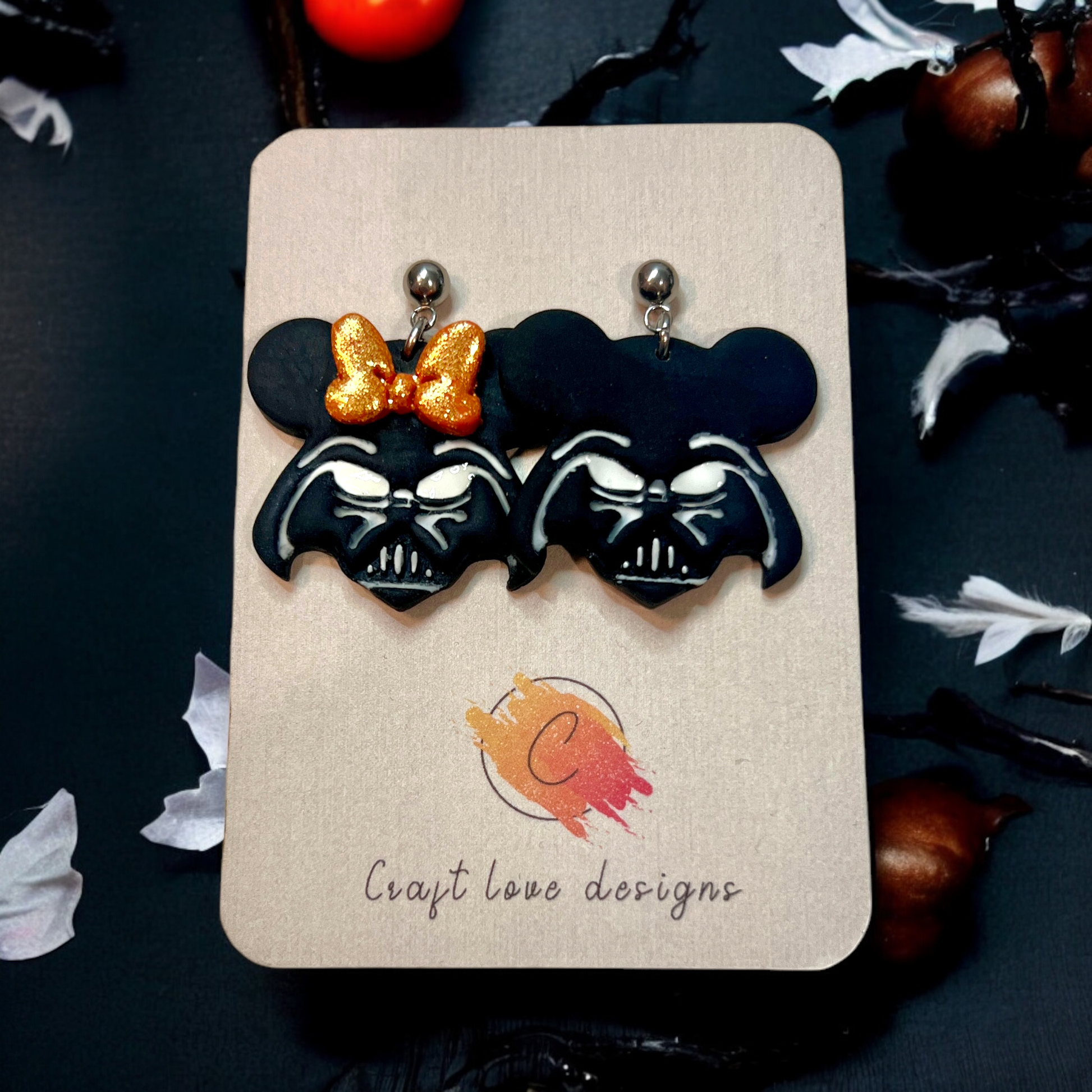 Black and glittery dark helmet mouse-themed earrings with a golden bow on a Craft Love Designs display card. Fun, bold accessory.