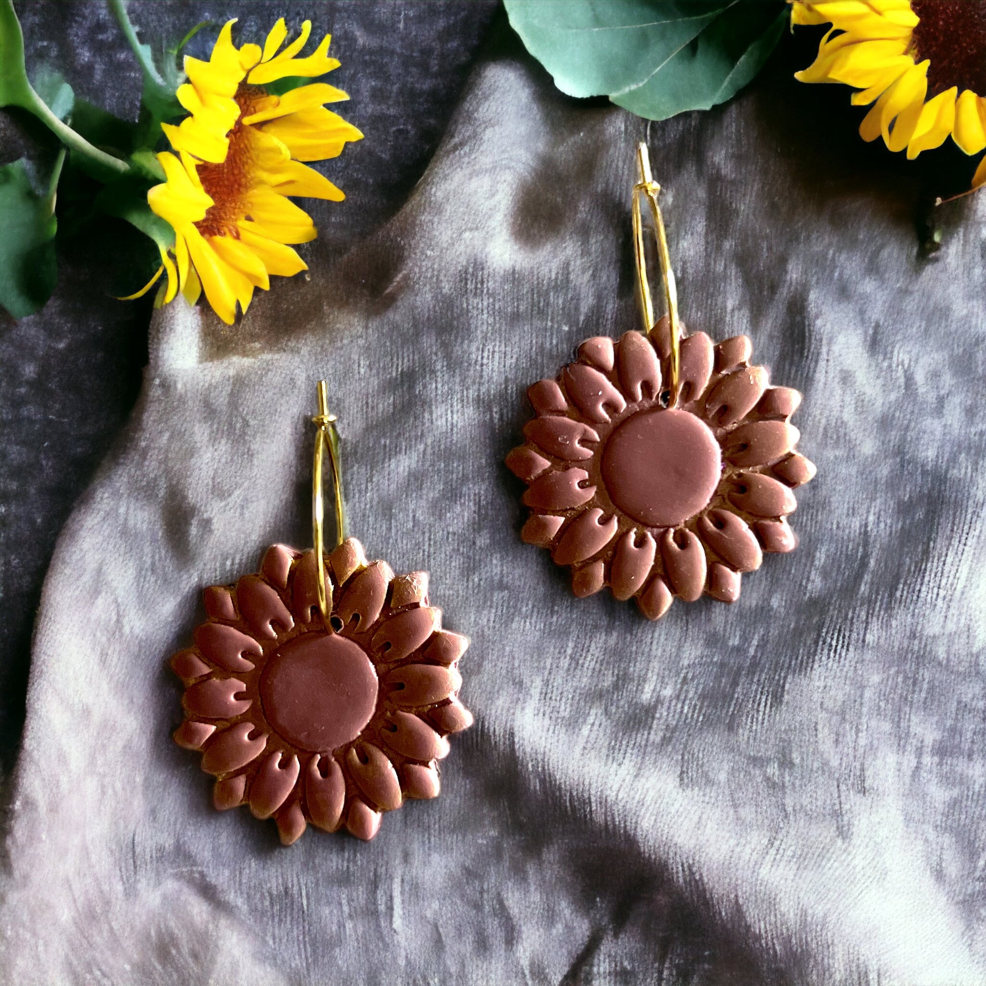 Dusty rose sunflower hoop earrings with textured petals and gold hoops, beautifully displayed with sunflowers in the background. 