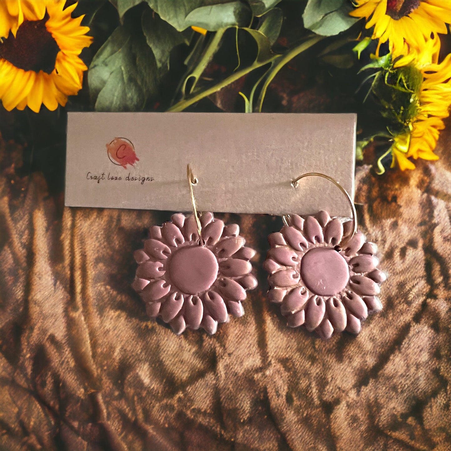 Close-up of dusty rose sunflower earrings on gold hoops, arranged on soft fabric with bright yellow sunflowers in the background.
