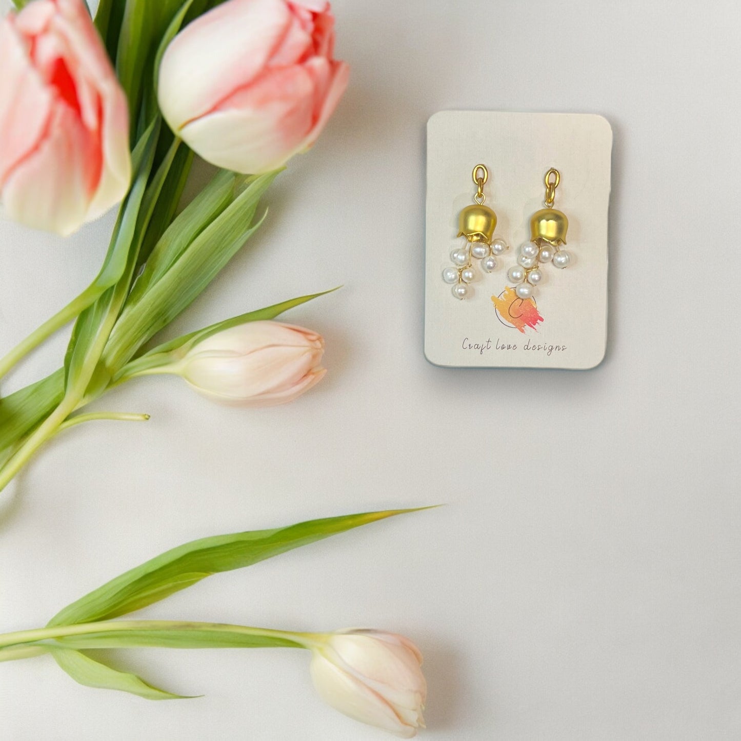 Elegant gold bellflower earrings with cascading pearls, displayed on a branded Craft Love Designs card and surrounded by pink tulips.