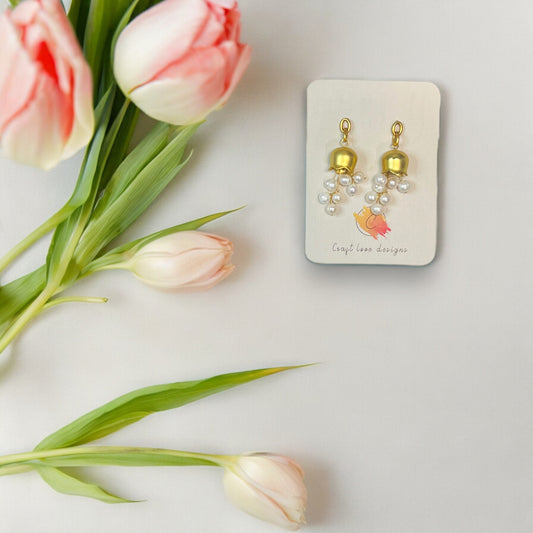 Elegant gold bellflower earrings with cascading pearls, displayed on a branded Craft Love Designs card and surrounded by pink tulips.