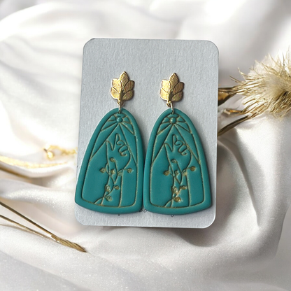 Elegant teal earrings with embossed floral patterns and gold leaf studs displayed on a Craft Love Designs card against white silk fabric.