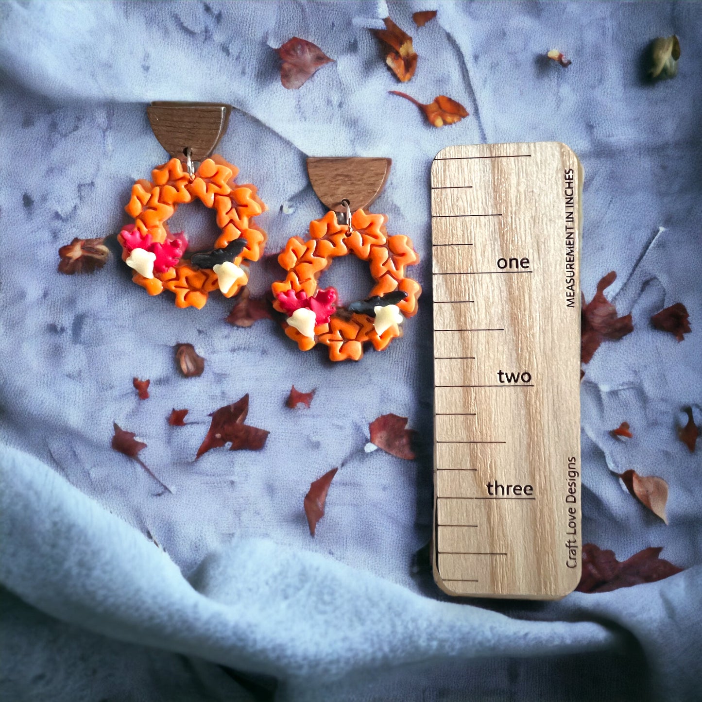 Orange leaf wreath earrings shown next to a size scale, surrounded by scattered autumn leaves on soft fabric.