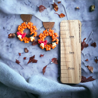 Orange leaf wreath earrings shown next to a size scale, surrounded by scattered autumn leaves on soft fabric.