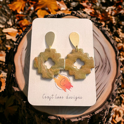 Beige and fall-inspired Aztec style dangle earrings with a unique cutout design, featuring golden teardrop tops, set against a tree bark with autumn leaves. 