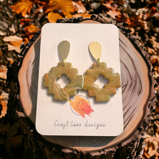 Beige and fall-inspired Aztec style dangle earrings with a unique cutout design, featuring golden teardrop tops, set against a tree bark with autumn leaves. 