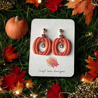 Handmade pumpkin-shaped dangle earrings with a sparkling crystal charm displayed on a branded card, surrounded by autumn leaves and decor.