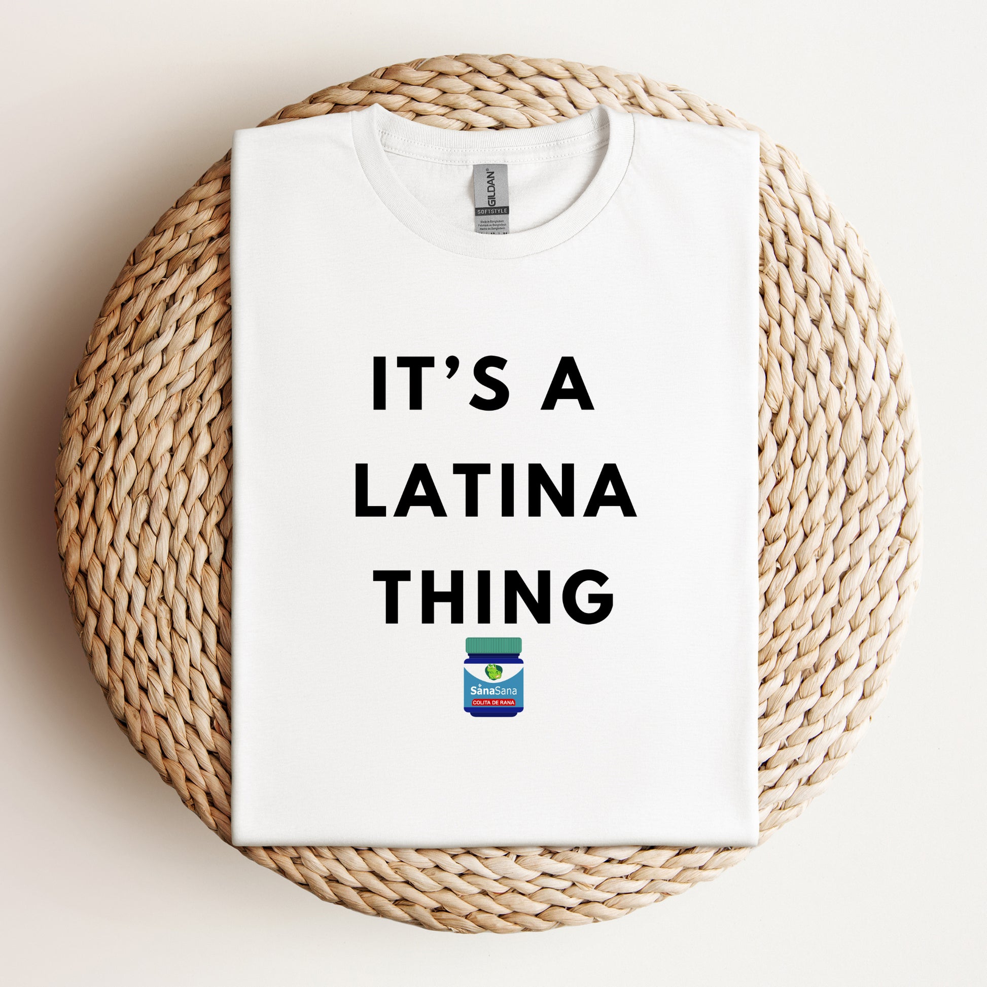 Flat lay of a white t-shirt with bold black text reading "It's a Latina Thing" and a small Vicks-inspired design, displayed on a woven background.
