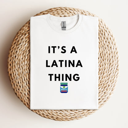 Flat lay of a white t-shirt with bold black text reading "It's a Latina Thing" and a small Vicks-inspired design, displayed on a woven background.