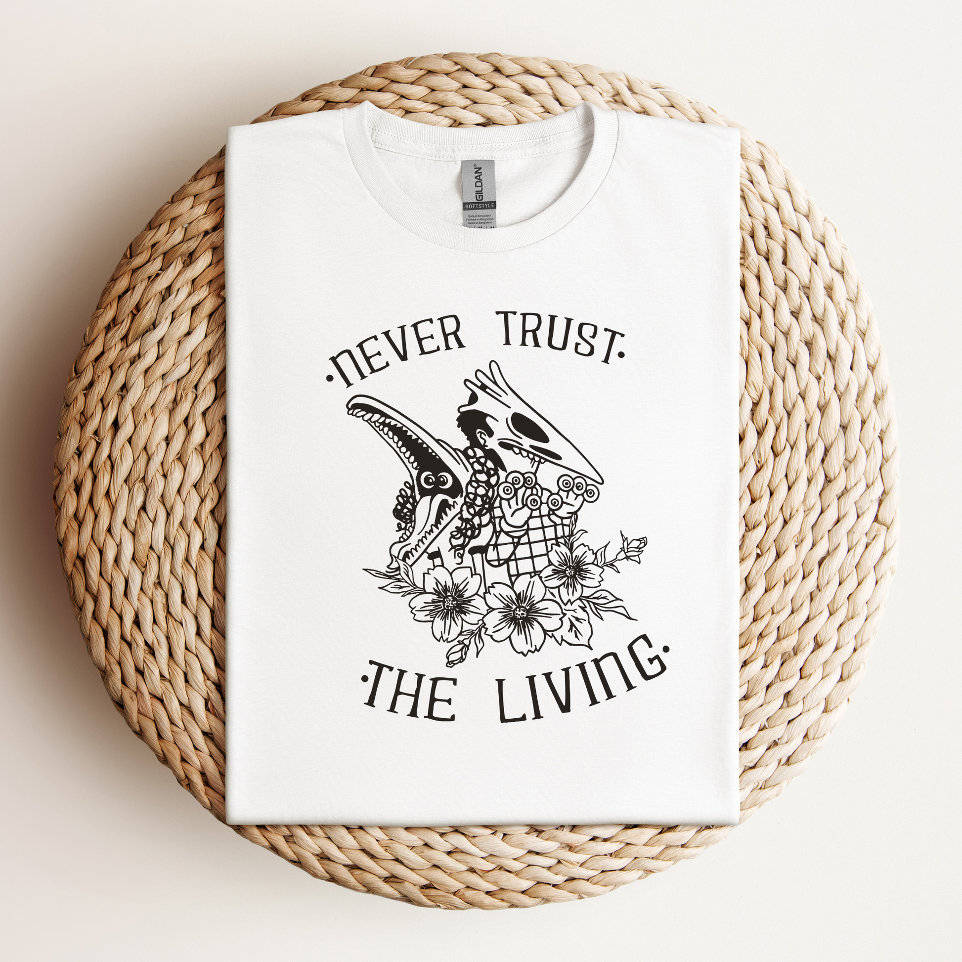 Flat lay of a white t-shirt with black gothic-style artwork featuring skulls, flowers, and the phrase "Never Trust the Living," displayed on a woven surface.