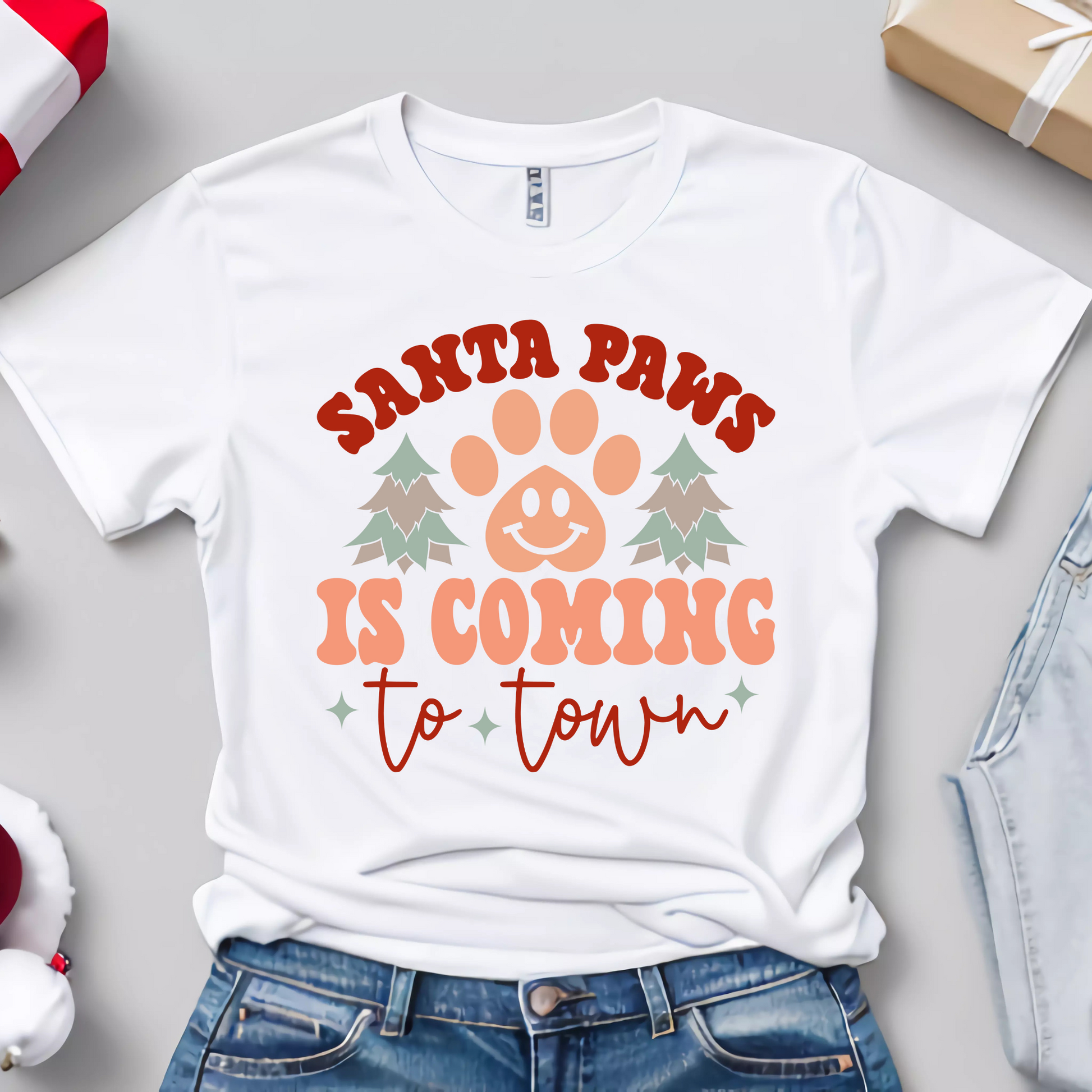 Flat lay of a white t-shirt featuring the text "Santa Paws Is Coming to Town" with a cute paw print, holiday trees, and retro-style design.