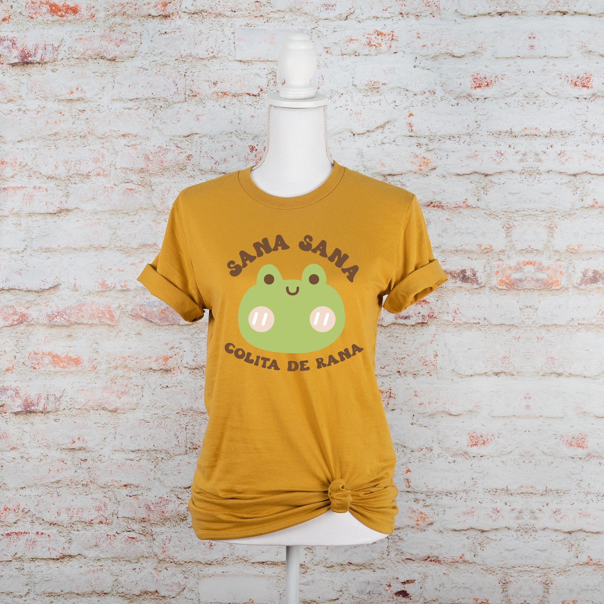 Flat lay of mustard yellow t-shirt with a green frog and Spanish phrase "Sana Sana Colita de Rana," placed on a neutral brick background.