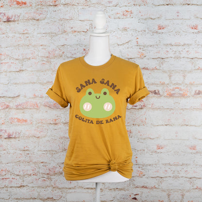 Flat lay of mustard yellow t-shirt with a green frog and Spanish phrase "Sana Sana Colita de Rana," placed on a neutral brick background.