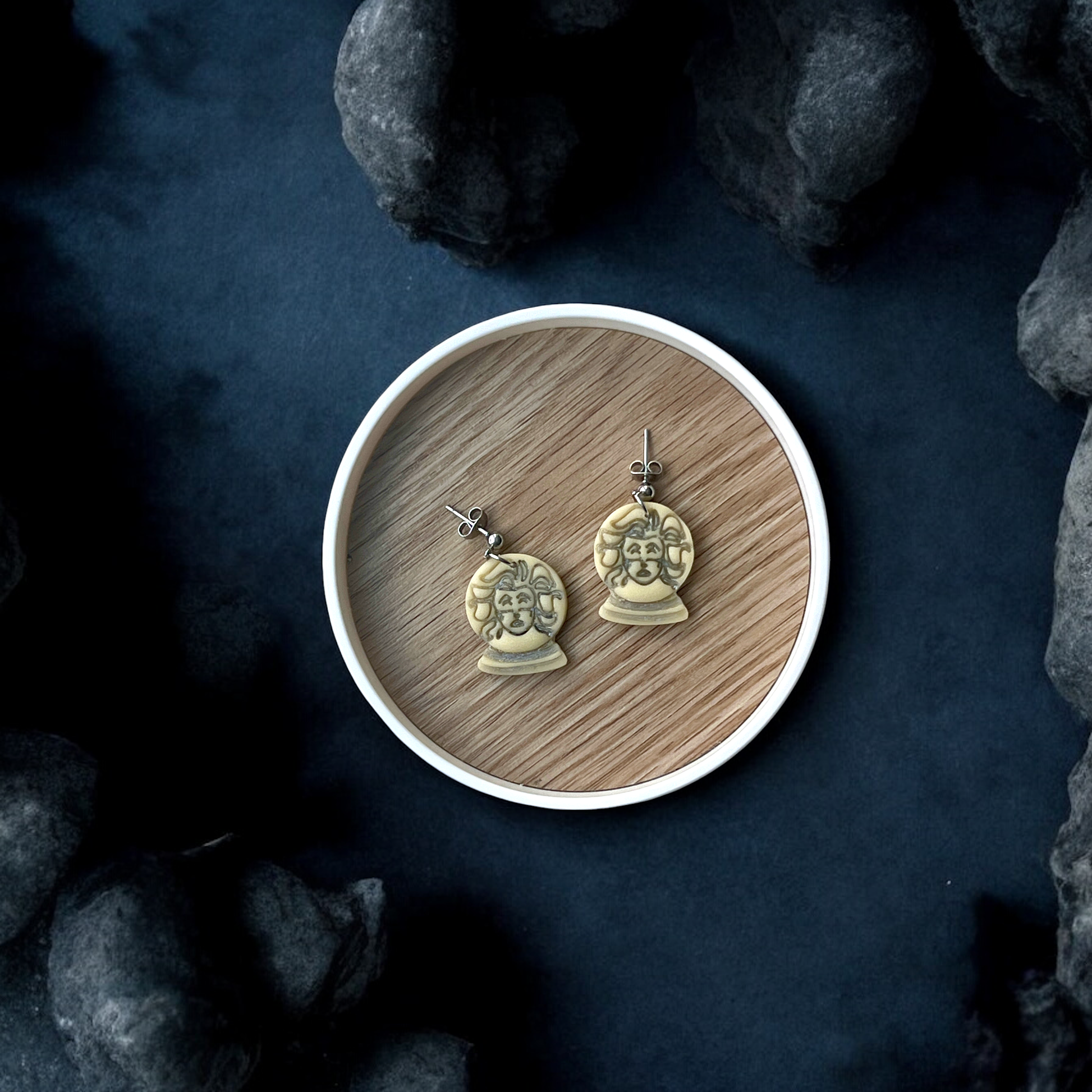 Pair of glow in the dark fortune teller bust earrings displayed inside a round wooden tray against a dark, mysterious background with stones. 