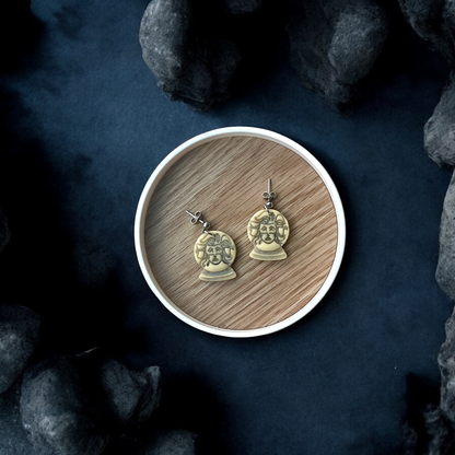 Pair of glow in the dark fortune teller bust earrings displayed inside a round wooden tray against a dark, mysterious background with stones. 