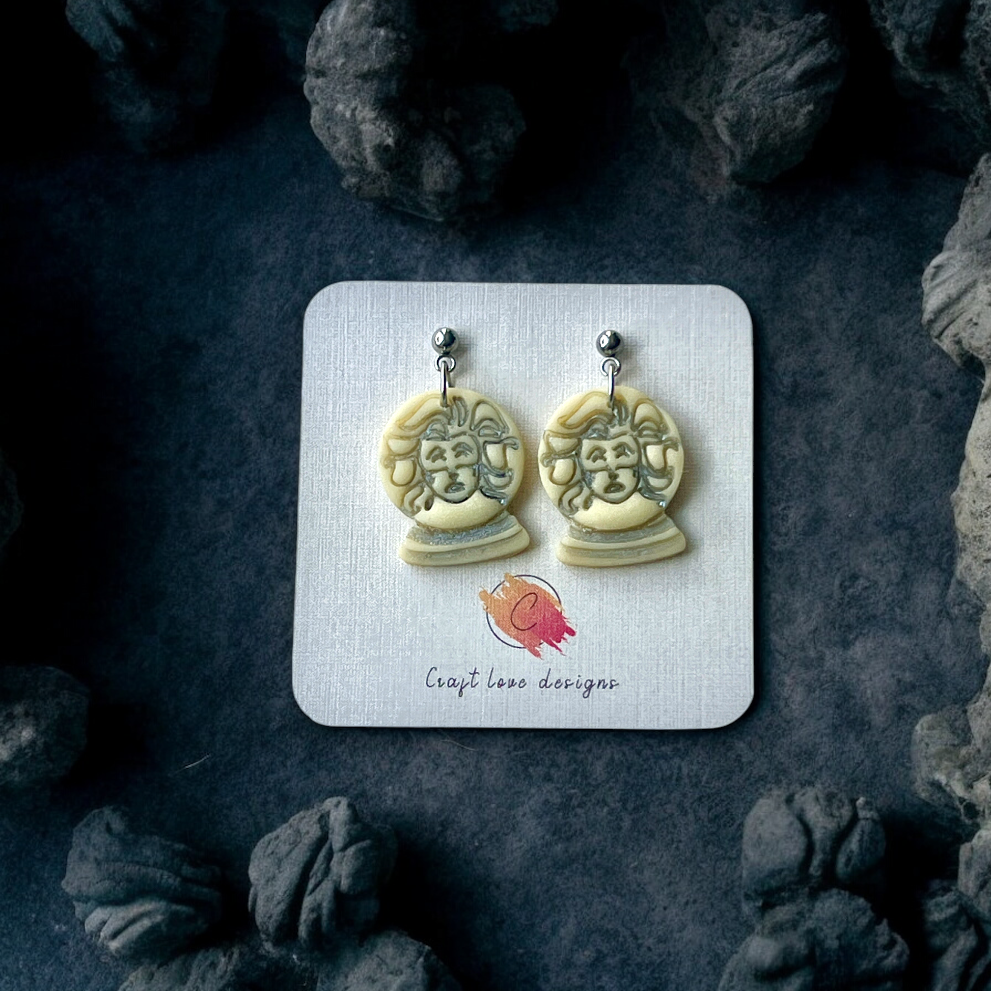 Glow in the dark fortune teller bust earrings showcased on a designer card, surrounded by eerie dark rocks, creating a spooky and mystical vibe.