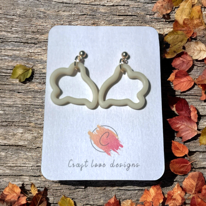 Cream-colored ghost-shaped earrings on a Craft Love Designs card, set against a rustic wooden background with scattered autumn leaves.