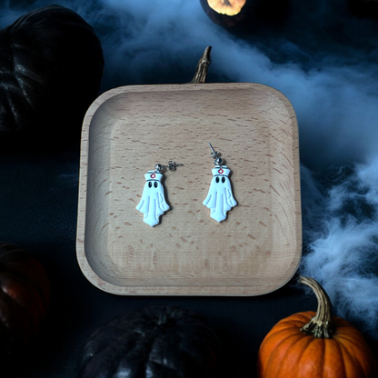 Ghost-themed earrings with nurse hats and a red cross, placed on a wooden tray surrounded by Halloween pumpkins and spooky fog effects. 
