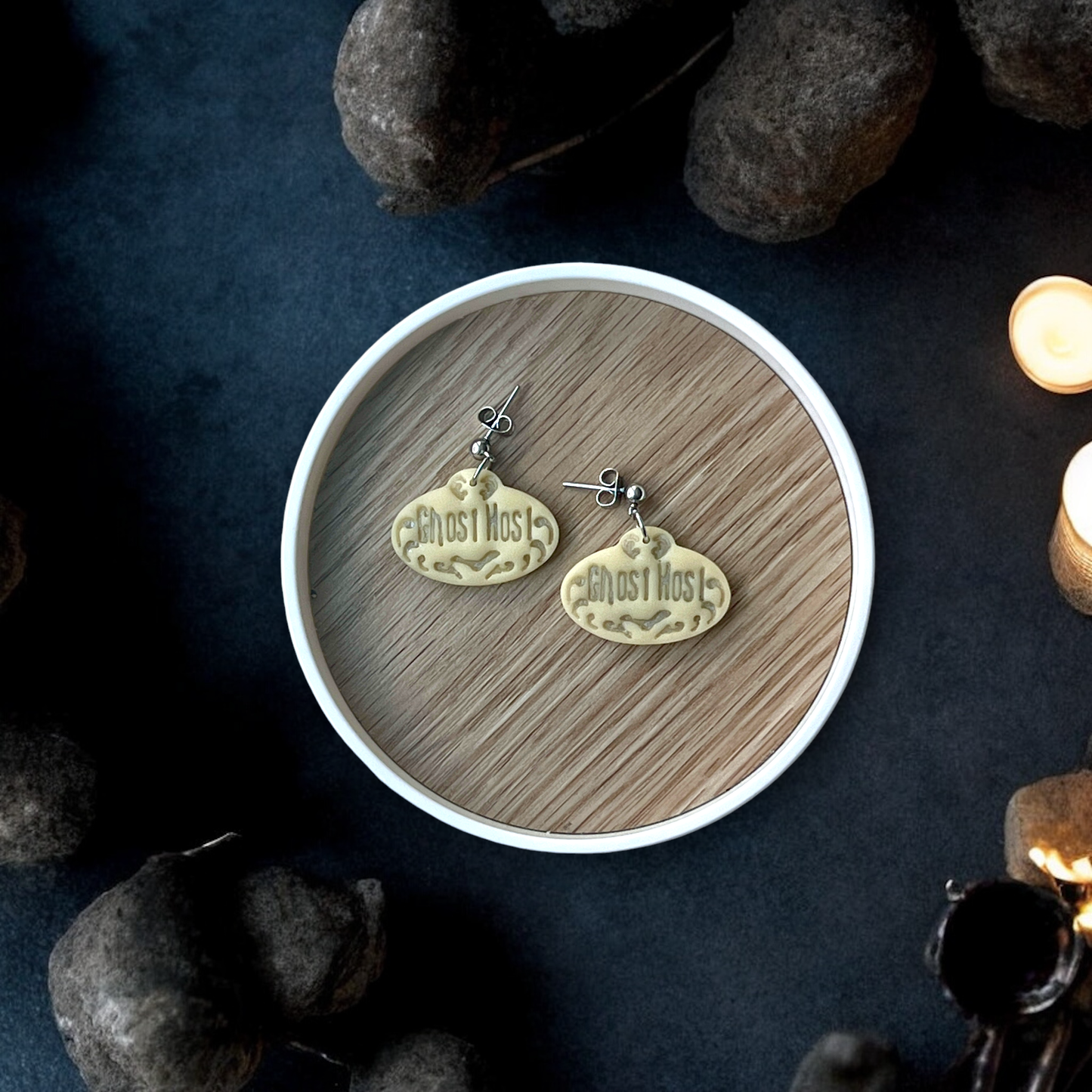 Spooky-themed "Ghost Host" earrings placed inside a round wooden tray with a moody dark background and eerie decor details nearby. 