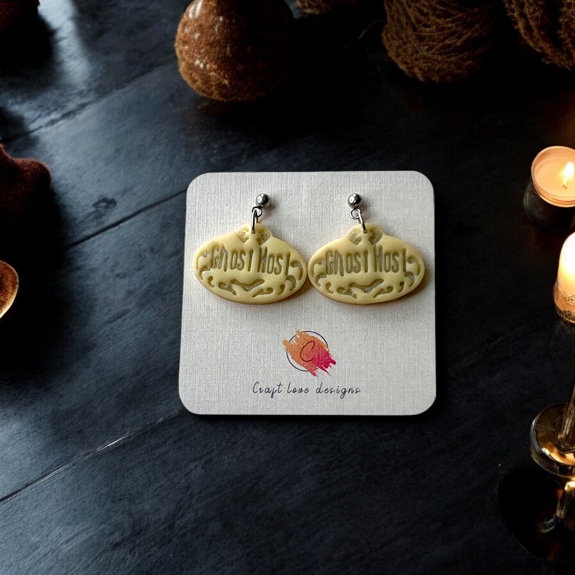Pair of glow in the dark "Ghost Host" earrings displayed on a designer card against a dark background with glowing candles and Halloween decor accents.