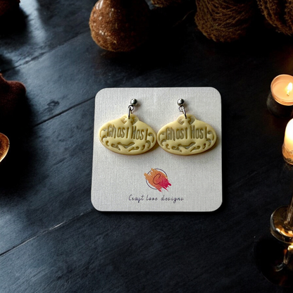 Pair of glow in the dark "Ghost Host" earrings displayed on a designer card against a dark background with glowing candles and Halloween decor accents.