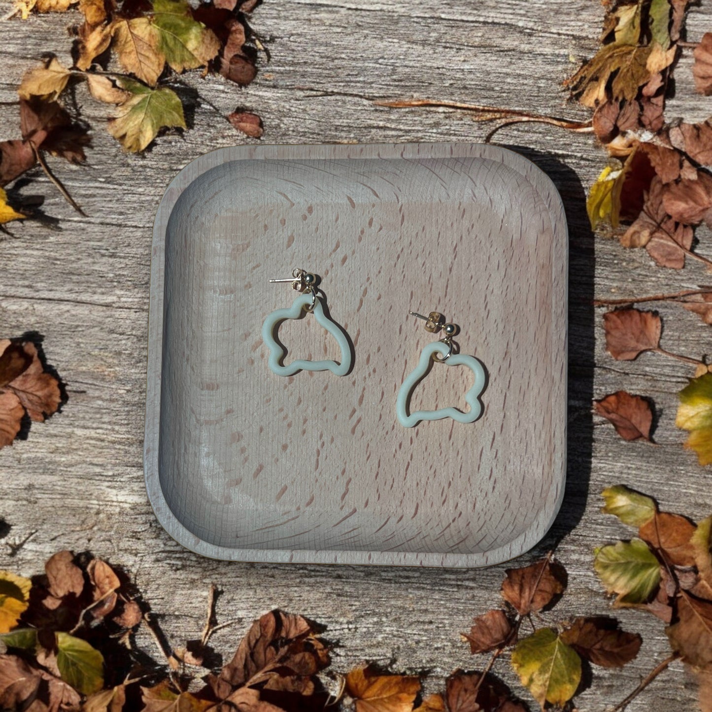 Cream ghost-shaped earrings resting in a wooden tray, placed on rustic wood, surrounded by scattered fall leaves for an autumn-inspired feel. 