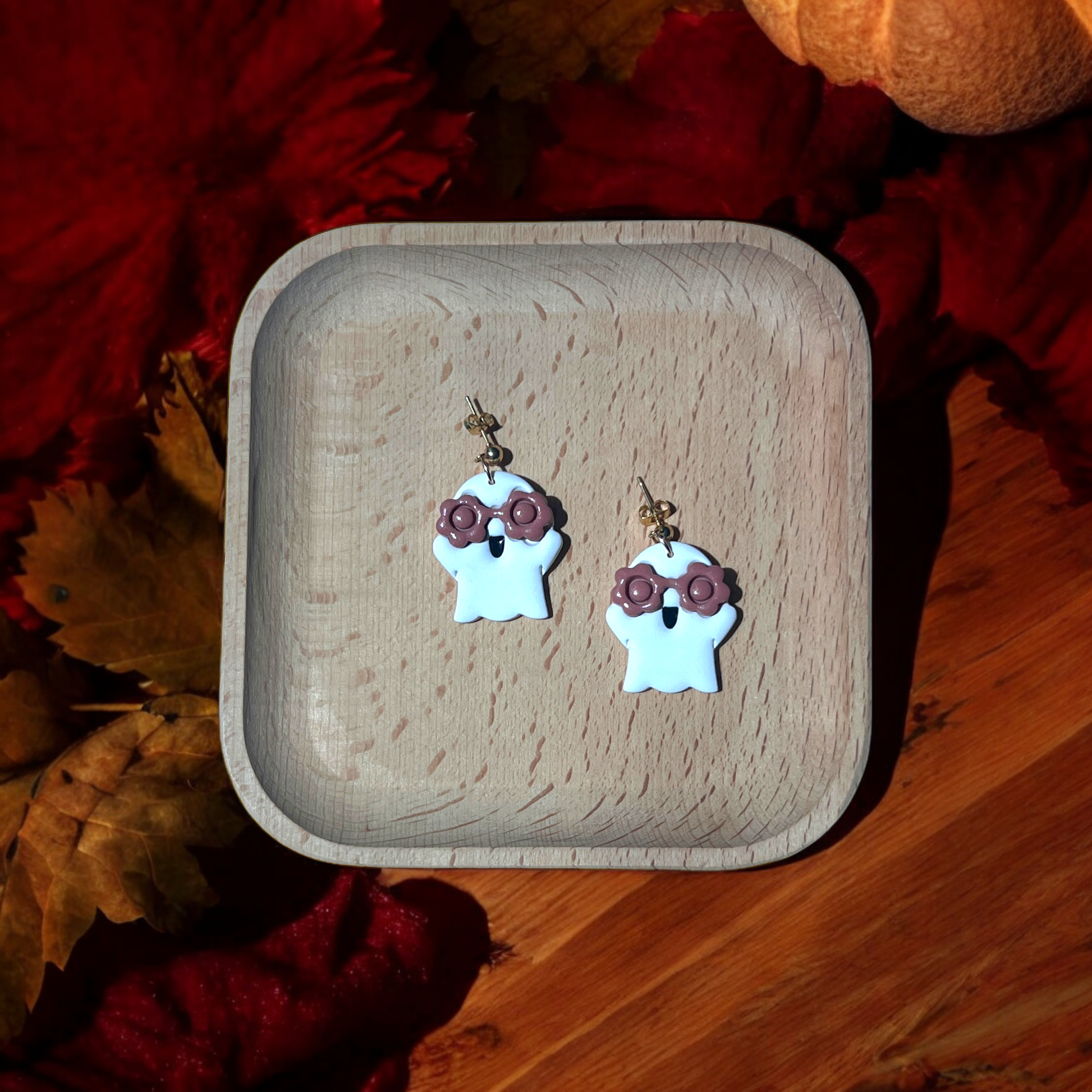 White ghost earrings with brown floral glasses resting in a wooden tray, surrounded by red autumn leaves and warm seasonal decor. 