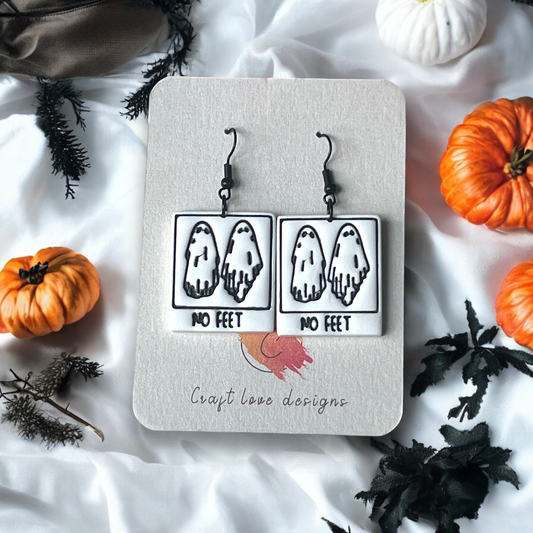 Black and white ghost earrings with "No Feet" text on a Craft Love Designs display card, surrounded by pumpkins and spooky decor.