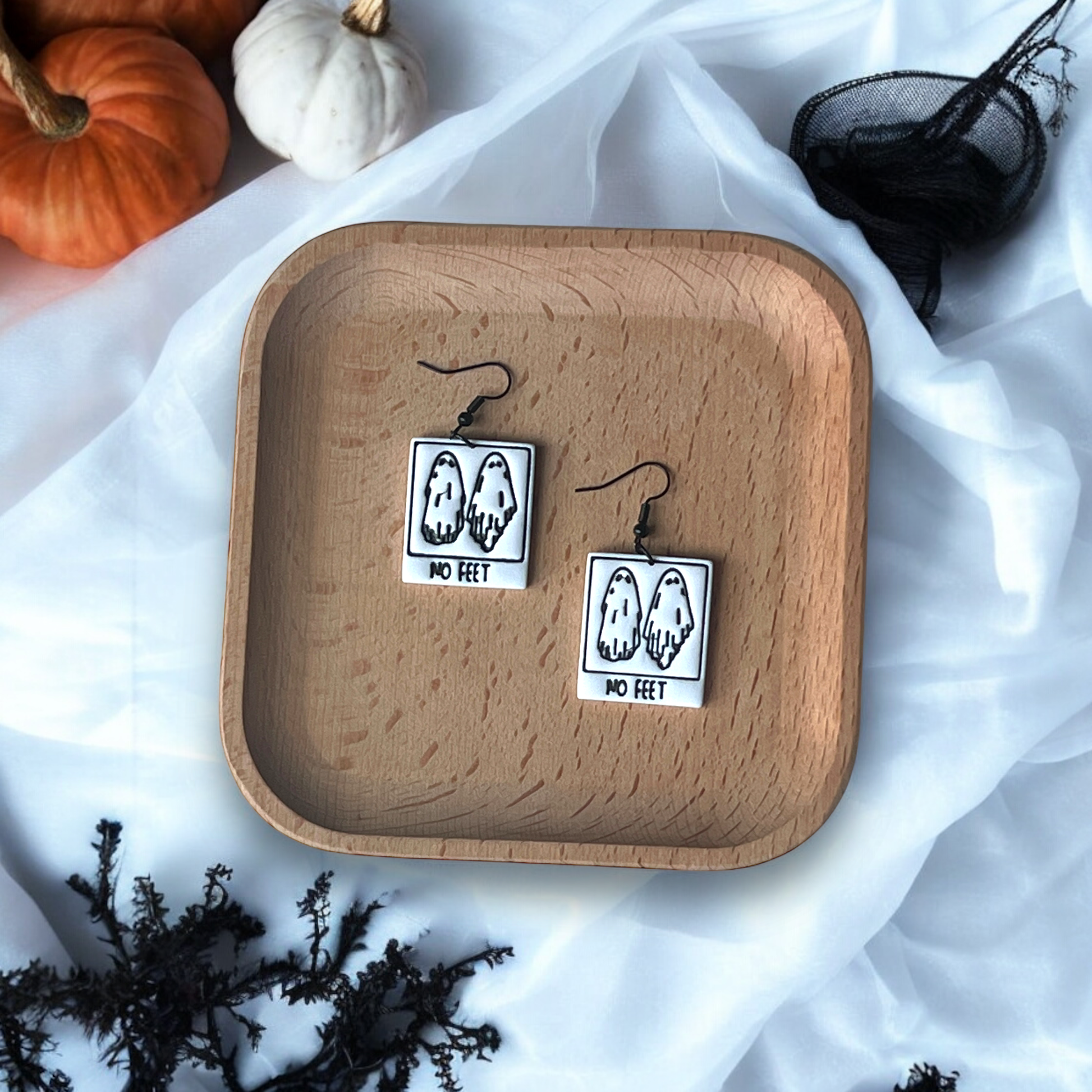 Black and white ghost earrings with "No Feet" text resting on a wooden tray, surrounded by pumpkins, branches, and a spooky setting. 