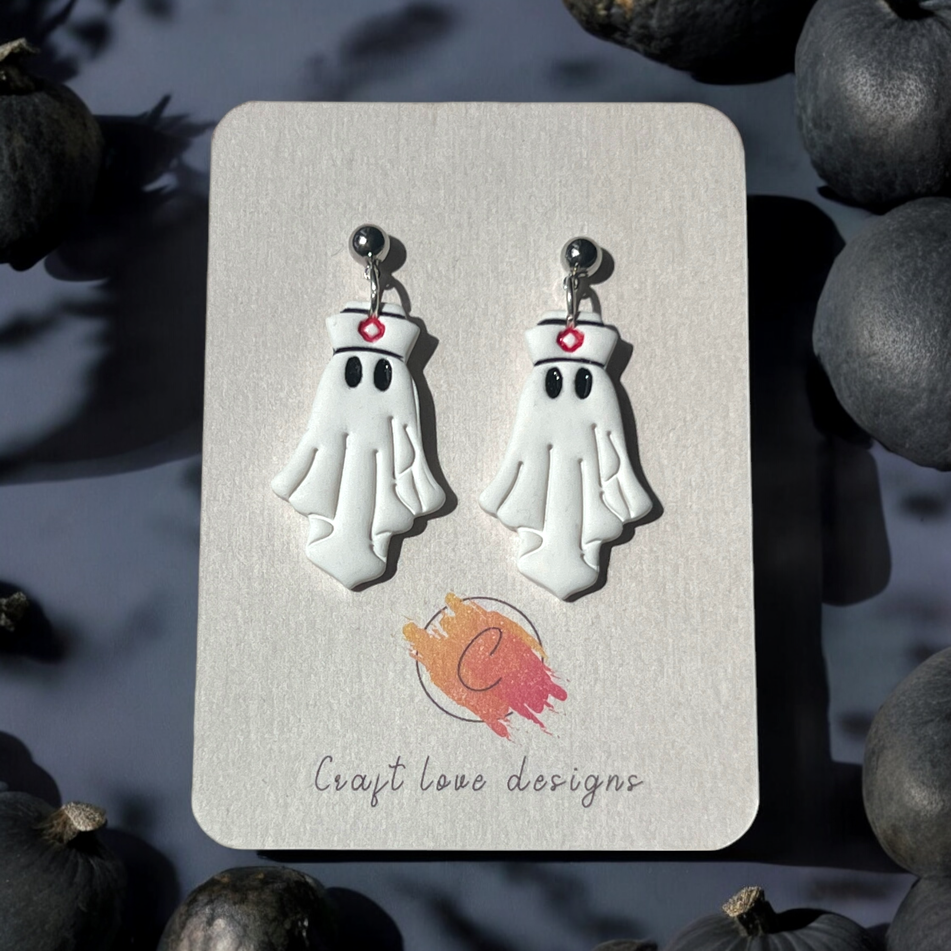 White ghost-shaped earrings featuring a nurse hat with a red cross, displayed on a branded card with dark pumpkins in the background.
