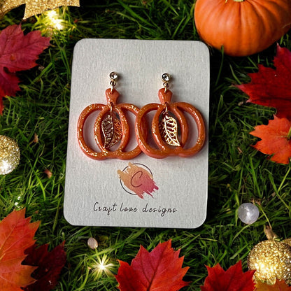 Glittery pumpkin-shaped dangle earrings with gold leaf accents displayed on a branded card, surrounded by fall leaves and festive lights.