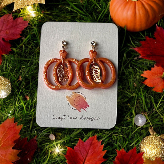 Glittery pumpkin-shaped dangle earrings with gold leaf accents displayed on a branded card, surrounded by fall leaves and festive lights.