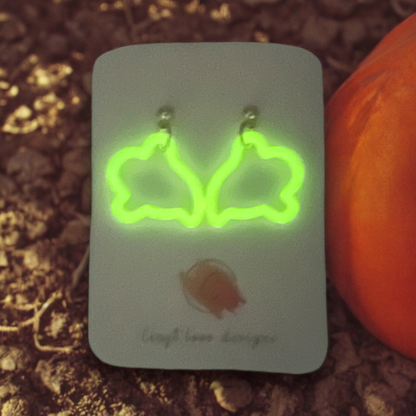 Bright green, glow-in-the-dark ghost-shaped earrings on a display card, set against dirt with a pumpkin for a spooky nighttime effect.