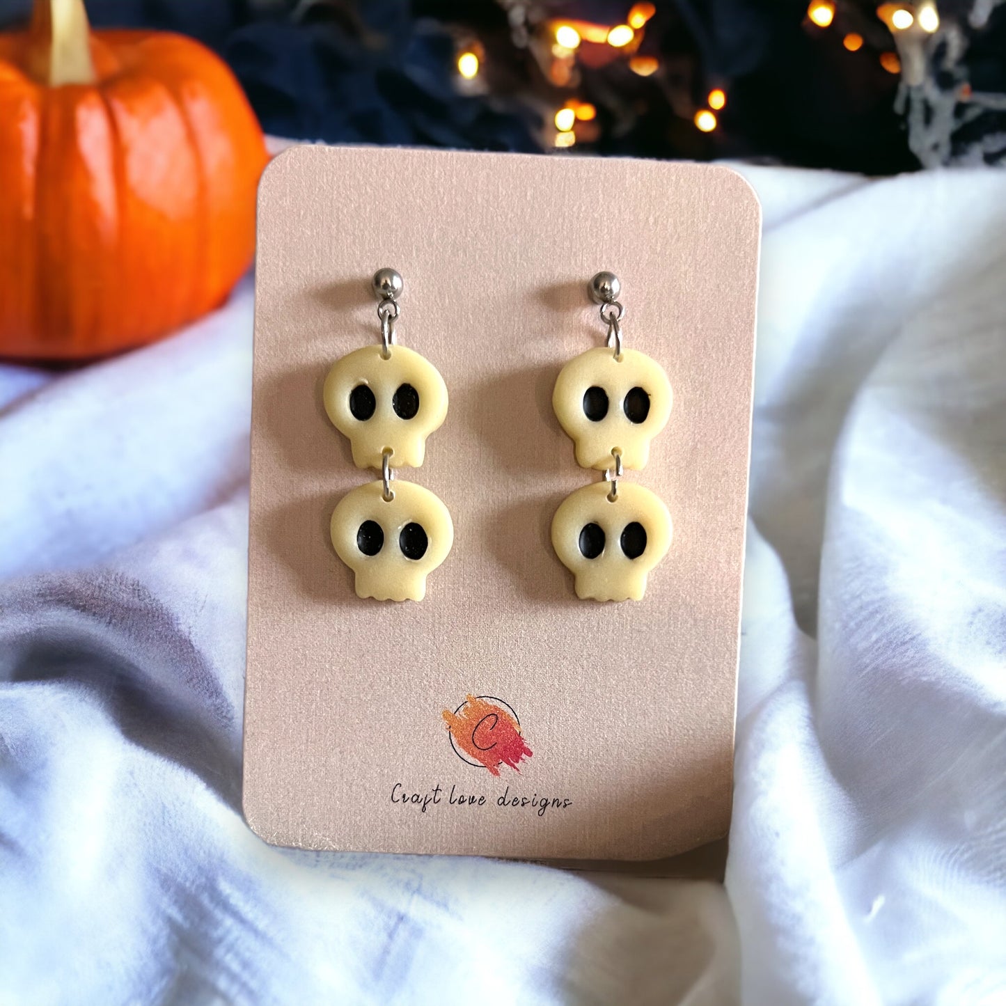 Close-up of white glow-in-the-dark skull dangle earrings on a beige card, with a festive pumpkin in the background. 