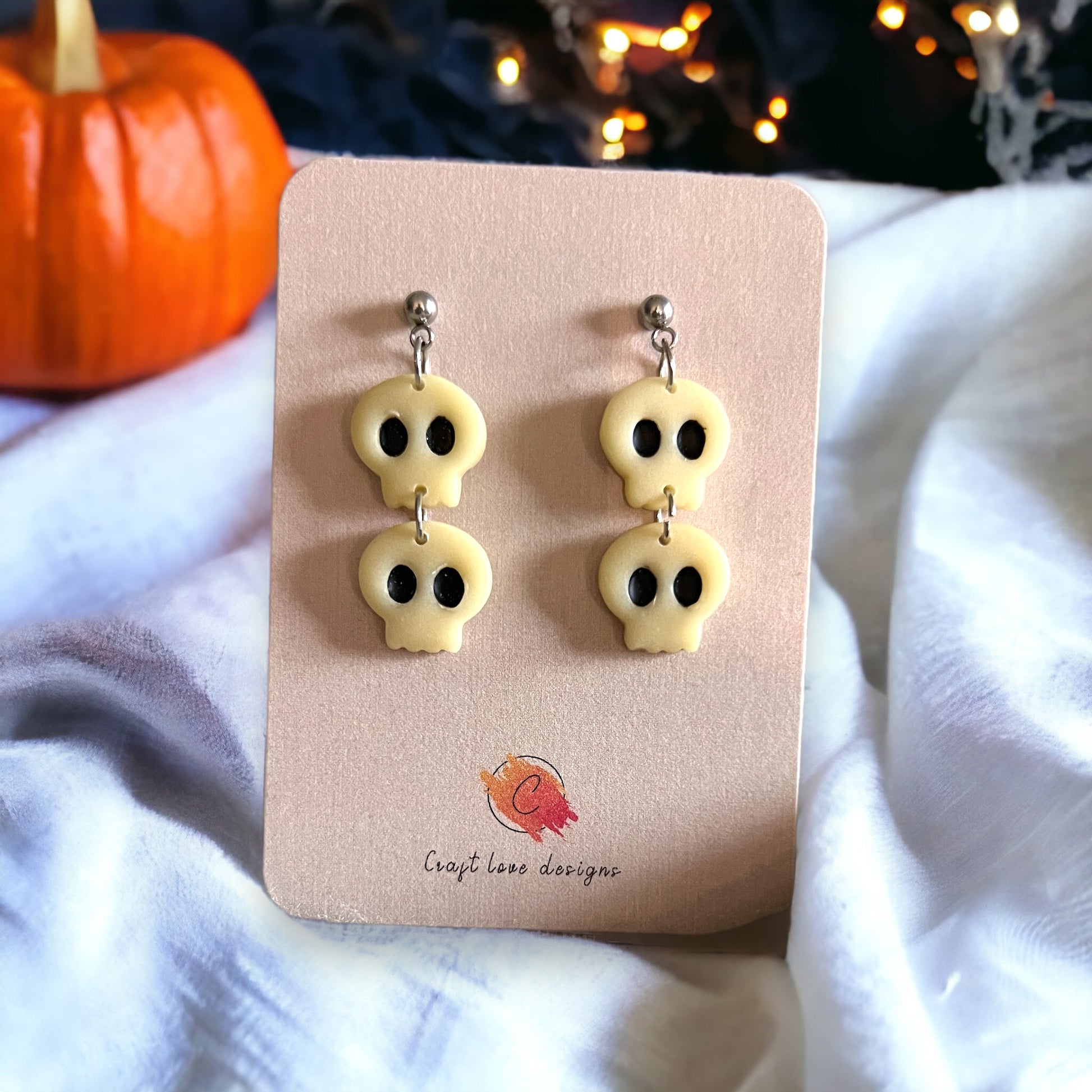 Close-up of white glow-in-the-dark skull dangle earrings on a beige card, with a festive pumpkin in the background. 