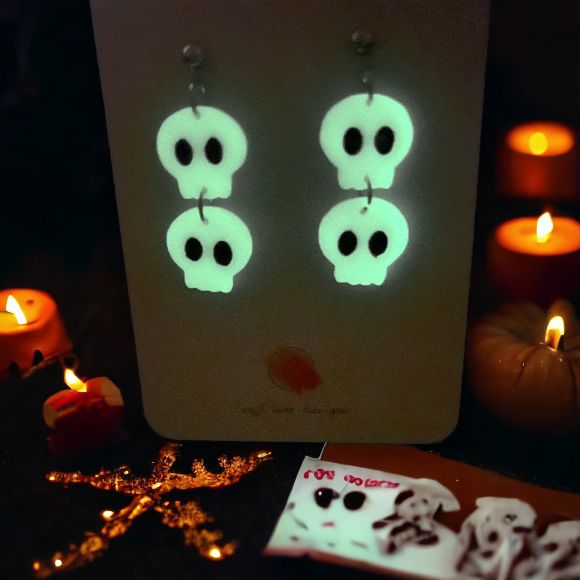 White skull-shaped dangle earrings glowing brightly in the dark, creating a spooky effect among Halloween props and candles.