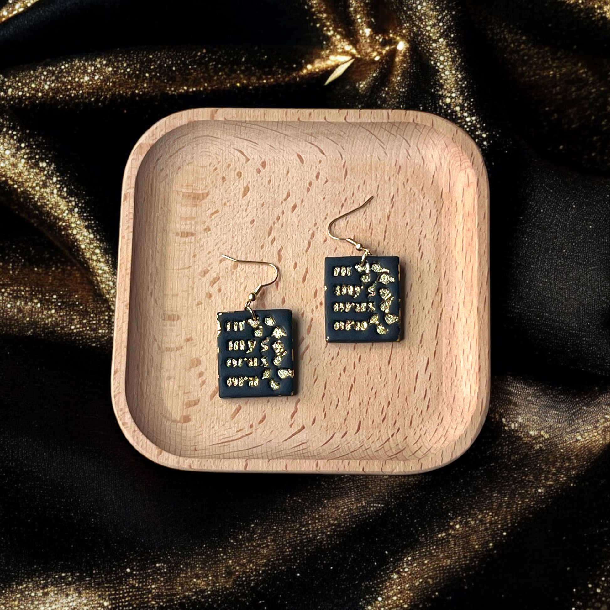 Handmade black square earrings with gold accents displayed in a wooden tray, placed on a shimmering gold fabric background