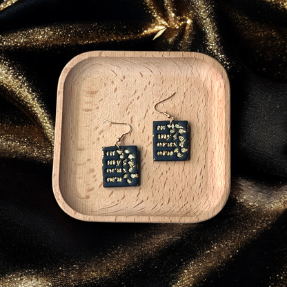 Handmade black square earrings with gold accents displayed in a wooden tray, placed on a shimmering gold fabric background