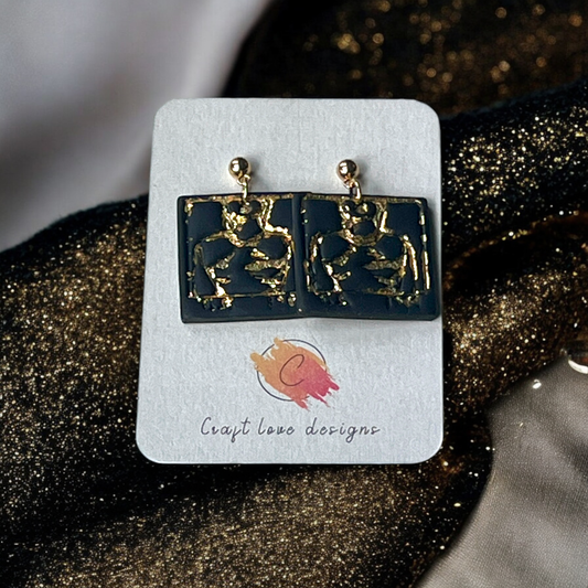 Square black drop earrings with textured abstract gold detailing, mounted on a display card, set against a backdrop of gold and black fabric. 