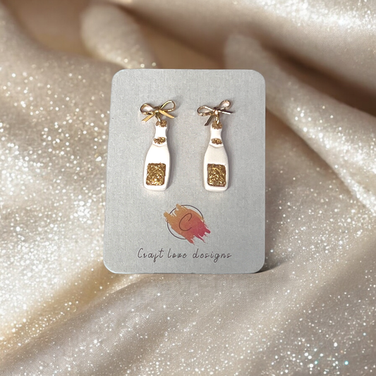 Gold-accented champagne bottle earrings with bow studs displayed on a Craft Love Designs card, set against shimmering fabric.