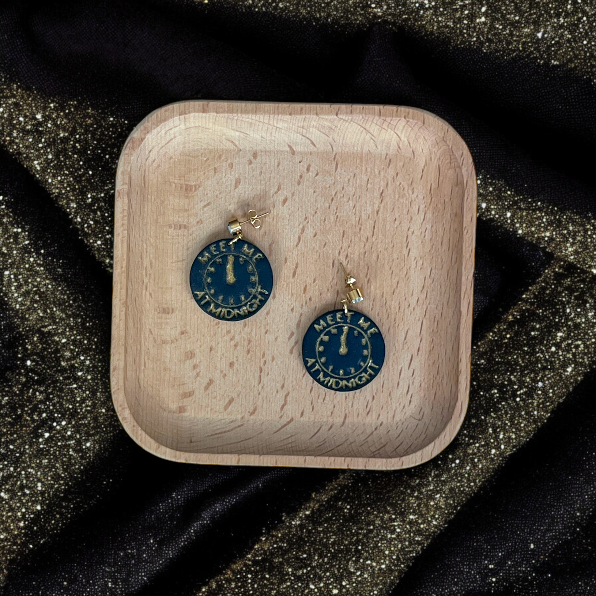Circular earrings displaying a gold clock striking midnight with engraved text, placed on a wooden tray surrounded by shimmering black and gold fabric.
