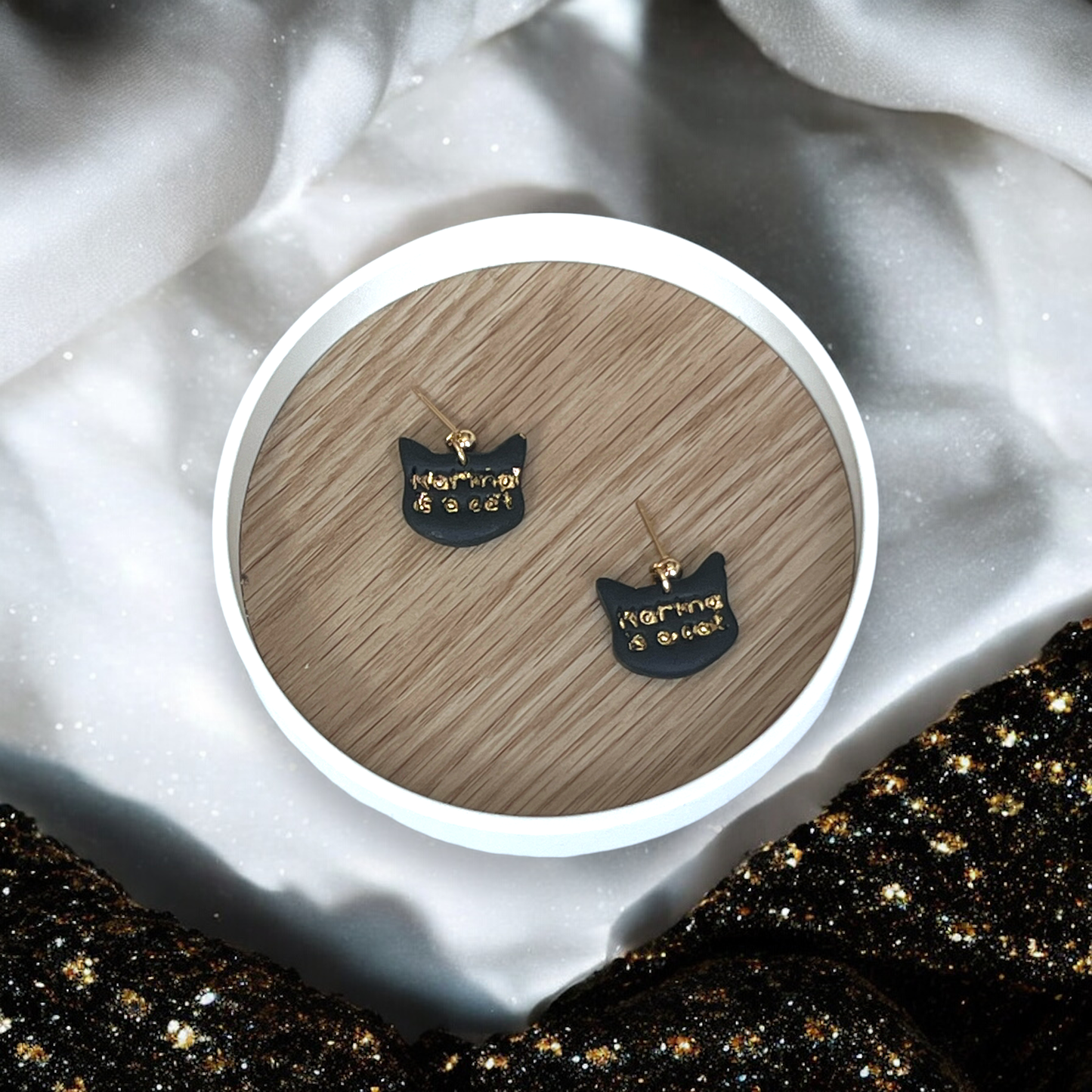 Black cat-shaped earrings with gold-embossed "Karma is a Cat" text, resting on a wooden tray with soft, glittery fabric surroundings.